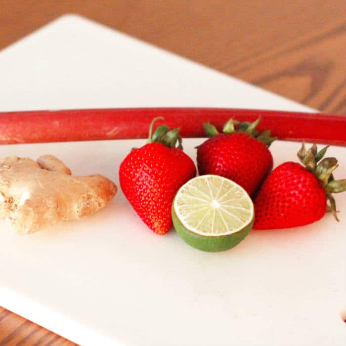 stalk of rhubarb, knob of ginger, strawberries, and a lime on a white cutting board