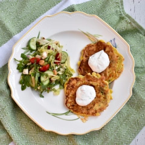 two gluten free zucchini pancakes topped with yogurt sauce on serving plate with a side salad